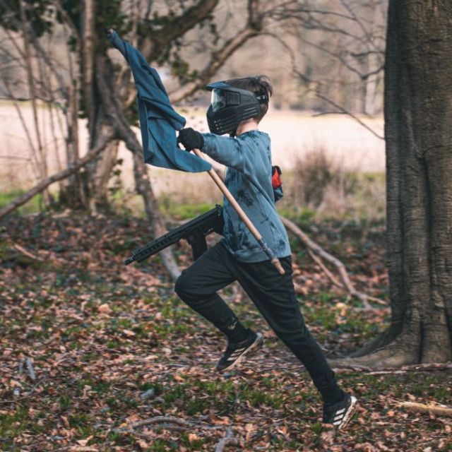 Person running in woods wearing a helmet, holding a blue flag and marker gun.