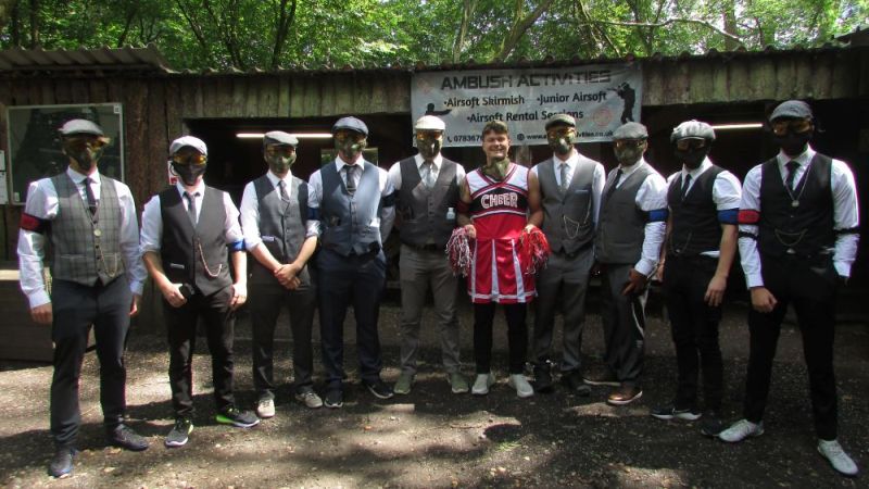 Group of men in suits and masks with one in a cheerleader outfit at an outdoor venue.