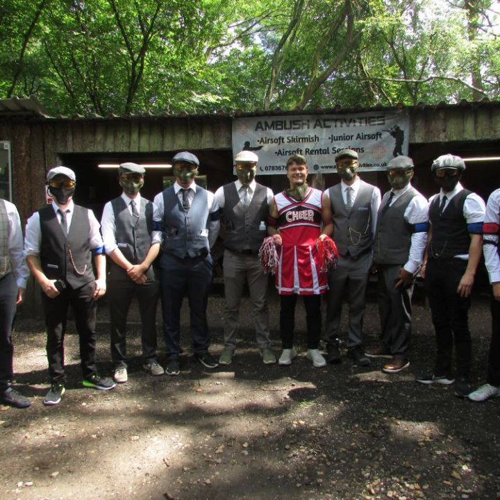 Group of men in suits and masks with one in a cheerleader outfit at an outdoor venue.
