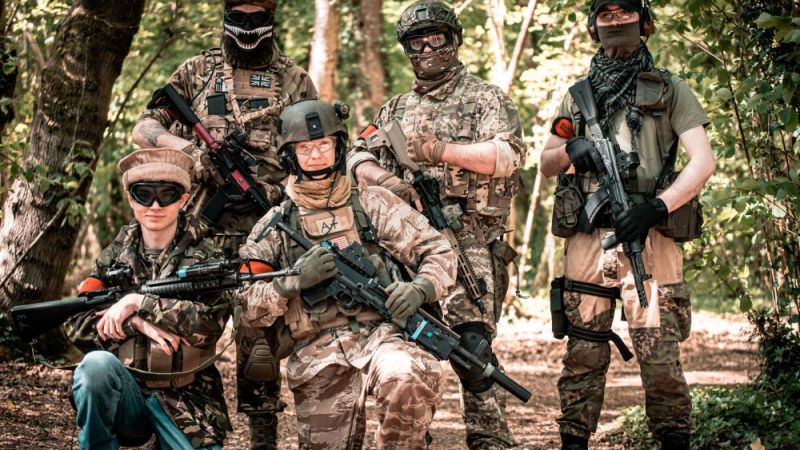 Five people in camouflage gear and helmets holding rifles in a forest setting.